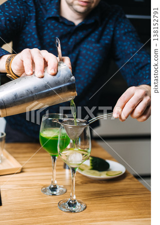 Mixing glass containing green liquid and cucumber slices sits on wooden counter next to metal shaker and lime. home bartending, craft green cocktails, mixology trend, DIY cocktail recipes Mixing glass containing green liquid and cucumber slices sits on wooden counter next to metal shaker and lime. home bartending, craft green cocktails, mixology trend, DIY cocktail recipes 138152791