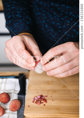 Hands peel fresh lychee fruit over wooden board next to knife and cloth. Lychee fruit, tropical ingredients, botanical ingredients, gourmet ingredients, culinary innovation. 138152808