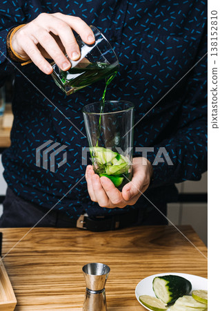 Hands pour green liquid into glass filled with cucumber slices on wooden counter. cucumber mint fizz, refreshing cucumber mocktail, hydrating gin drinks, cucumber ribbon garnish Hands pour green liquid into glass filled with cucumber slices on wooden counter. cucumber mint fizz, refreshing cucumber mocktail, hydrating gin drinks, cucumber ribbon garnish 138152810