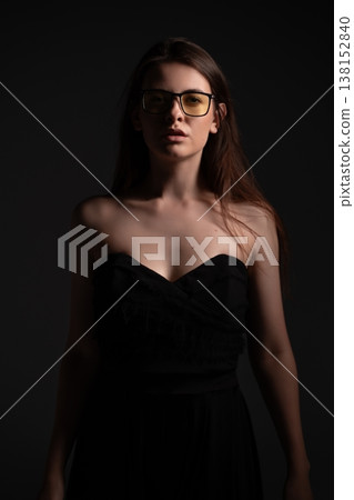 Young woman with glasses wearing a black strapless dress in low light, standing against a dark background, portraying elegance, mystery, and a confident mood in a fashion studio portrait 138152840