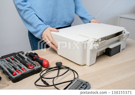 Hands position a white all-in-one printer on a desk with a toolkit and power cable, depicting DIY printer repair, office equipment maintenance and troubleshooting in a home or small-business workspace Hands position a white all-in-one printer on a desk with a toolkit and power cable, depicting DIY printer repair, office equipment maintenance and troubleshooting in a home or small-business workspace 138155371