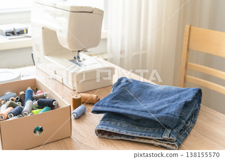 Sewing machine on wooden table beside folded blue jeans and an open box of colorful thread spools, sunlight from window illuminating workspace 138155570