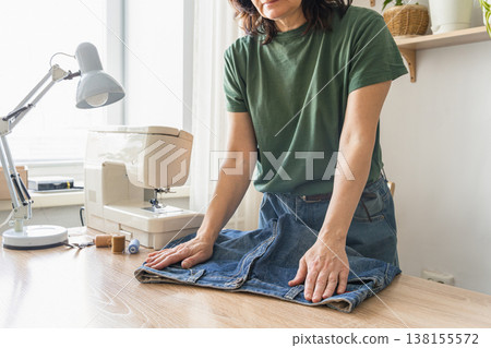 Woman standing at a desk, smoothing out a pair of jeans, preparing for sewing or alteration with a sewing machine nearby Woman standing at a desk, smoothing out a pair of jeans, preparing for sewing or alteration with a sewing machine nearby 138155572