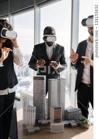 Three professionals in suits wearing VR headsets interact with a miniature city model during a virtual reality business meeting 138155632