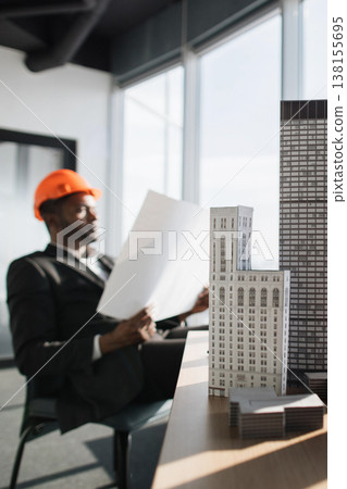 An architect wearing an orange hard hat reviews blueprints while sitting in an office with architectural models on a desk An architect wearing an orange hard hat reviews blueprints while sitting in an office with architectural models on a desk 138155695