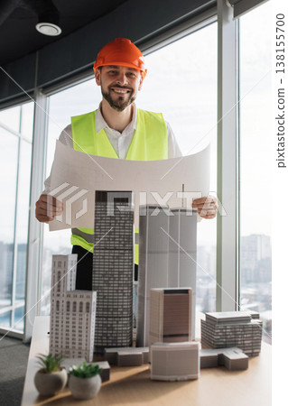 A smiling architect in a hard hat and safety vest holds blueprints while standing in front of a table with miniature building models A smiling architect in a hard hat and safety vest holds blueprints while standing in front of a table with miniature building models 138155700