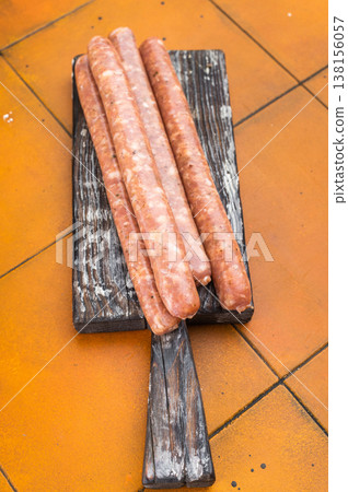 Raw Chipolata sausages with thyme and garlic arranged on wooden board with carving fork. Fresh seasoned pork meat on orange tile background for cooking concept. Raw Chipolata sausages with thyme and garlic arranged on wooden board with carving fork. Fresh seasoned pork meat on orange tile background for cooking concept. 138156057