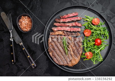 Juicy sliced ribeye steak with rich marbling and browned surface. Grilled rib eye beef served with salad on black ceramic plate. 138156292