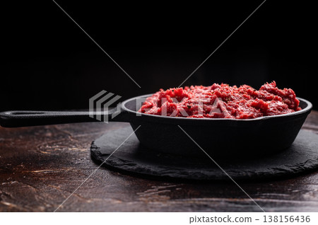 Raw ground moose game meat in a cast iron skillet on a black slate, set against a dark rustic brown background. Copy space. 138156436