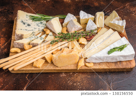 Brie Camembert Parmesan and Roquefort arranged with nuts herbs and breadsticks on a rustic wooden board over a dark textured background. 138156550