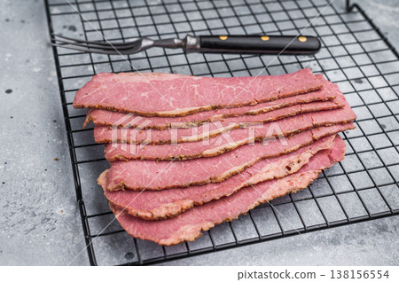 Thin sliced pastrami arranged on a metal cooling rack with a fork on a gray textured background. Smoked deli beef with marbled texture, ready for cooking and sandwiches. Thin sliced pastrami arranged on a metal cooling rack with a fork on a gray textured background. Smoked deli beef with marbled texture, ready for cooking and sandwiches. 138156554
