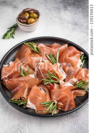 Delicious dry cured Jamon pork meat served on a black dish. Horizontal composition with lots of grey background. Delicious dry cured Jamon pork meat served on a black dish. Horizontal composition with lots of grey background. 138156613