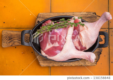 Raw duck legs in a black cast iron skillet, garnished with fresh thyme, resting on a rustic wooden cutting board against an orange tiled background. 138156648