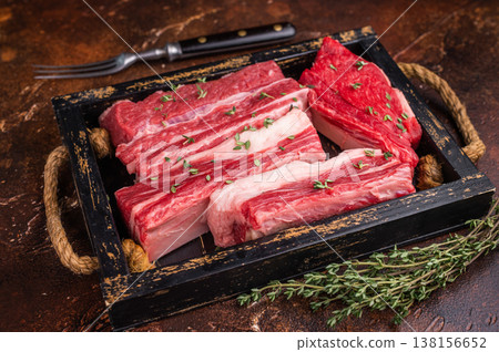 Raw beef short ribs with thyme and fork on a rustic dark background. Excellent for menu design or a butcher shop advertisement. Raw beef short ribs with thyme and fork on a rustic dark background. Excellent for menu design or a butcher shop advertisement. 138156652