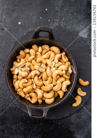 Golden cashew nuts displayed in a cast iron pan set on a slate board against a textured black surface giving a warm rustic look for recipes and snacks Golden cashew nuts displayed in a cast iron pan set on a slate board against a textured black surface giving a warm rustic look for recipes and snacks 138156702
