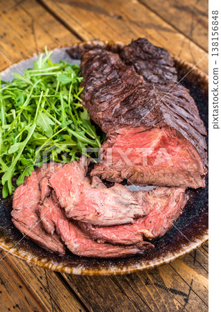 Closeup grilled onglet hanger steak with arugula salad on serving plate, seared crust and tender interior on rustic wood background Closeup grilled onglet hanger steak with arugula salad on serving plate, seared crust and tender interior on rustic wood background 138156848