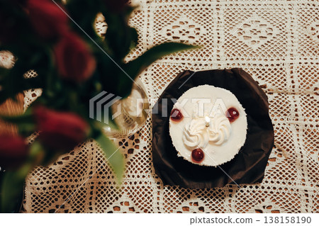 Overhead view of a small white cake with a candle on a lace tablecloth next to red roses. Birthday, anniversary celebration design themes. Overhead view of a small white cake with a candle on a lace tablecloth next to red roses. Birthday, anniversary celebration design themes. 138158190