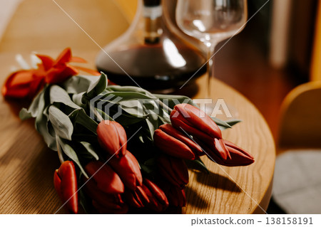 A bouquet of red tulips on a wooden table with a wine decanter and glass in the background. Moody vintage still life with soft lighting and film grain. Ideal for romantic and celebration designs. A bouquet of red tulips on a wooden table with a wine decanter and glass in the background. Moody vintage still life with soft lighting and film grain. Ideal for romantic and celebration designs. 138158191