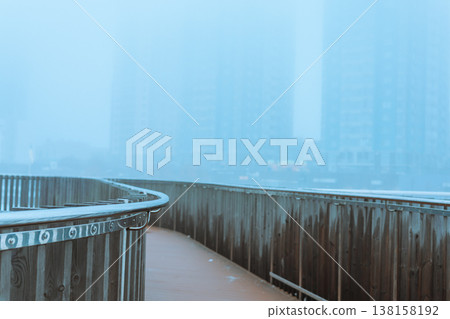 Urban landscape featuring a curved modern metal railing on a pedestrian walkway or bridge, skyscrapers in morning fog, mist. Atmospheric urban scene. Urban landscape featuring a curved modern metal railing on a pedestrian walkway or bridge, skyscrapers in morning fog, mist. Atmospheric urban scene. 138158192