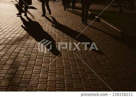Candid, high-contrast shot of people's silhouettes and long shadows walking on a paved urban sidewalk. The dramatic lighting creates an atmospheric effect, capturing a moment of everyday city life. Candid, high-contrast shot of people's silhouettes and long shadows walking on a paved urban sidewalk. The dramatic lighting creates an atmospheric effect, capturing a moment of everyday city life. 138158211