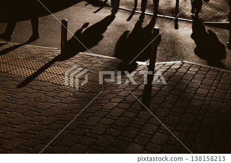 Candid, high-contrast shot of people's silhouettes and long shadows walking on a paved urban sidewalk. The dramatic lighting creates an atmospheric effect, capturing a moment of everyday city life. Candid, high-contrast shot of people's silhouettes and long shadows walking on a paved urban sidewalk. The dramatic lighting creates an atmospheric effect, capturing a moment of everyday city life. 138158213