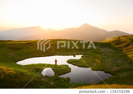 Small alpine lake reflecting sunrise light surrounded by green grass Small alpine lake reflecting sunrise light surrounded by green grass 138158296