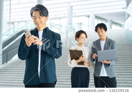 A middle-aged Japanese man smiles while operating his smartphone, while a business team holds a meeting in the background. A middle-aged Japanese man smiles while operating his smartphone, while a business team holds a meeting in the background. 138159516