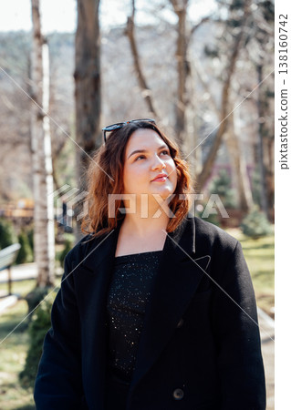 Thoughtful slavic appearance young woman in black coat standing in a zoo park looking up surrounded by trees and soft sunlight creating calm outdoor portrait Thoughtful slavic appearance young woman in black coat standing in a zoo park looking up surrounded by trees and soft sunlight creating calm outdoor portrait 138160742