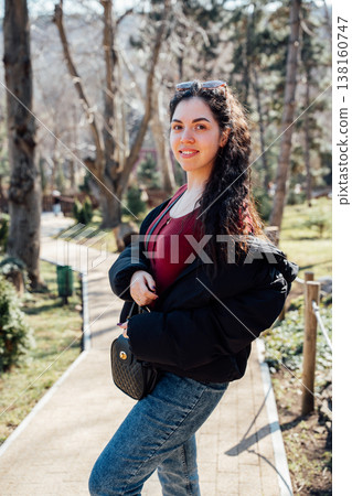 Smiling slavic appearance young woman walking in a zoo park wearing red sweater jeans and black jacket posing on a sunny path surrounded by trees 138160747