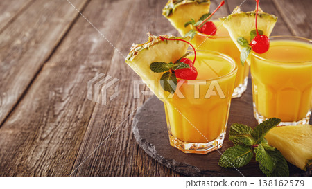 Glasses of pineapple juice with pieces of pineapple, cocktail cherry and mint on wooden table. Glasses of pineapple juice with pieces of pineapple, cocktail cherry and mint on wooden table. 138162579