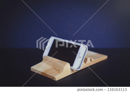 Horizontal perspective view of a handcrafted light wood smartphone stand with a wave-like carved surface, holding a white mobile phone against a dark blue background. 138163113