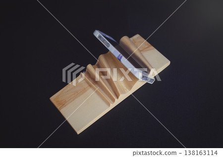 Horizontal high-angle view of a handcrafted light wood smartphone stand with a wave-like carved surface, holding a white mobile phone. 138163114