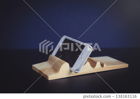 Horizontal close-up of a handcrafted light wood phone stand with a carved wave surface, securely holding a white smartphone at an angle against a dark blue background. Horizontal close-up of a handcrafted light wood phone stand with a carved wave surface, securely holding a white smartphone at an angle against a dark blue background. 138163116