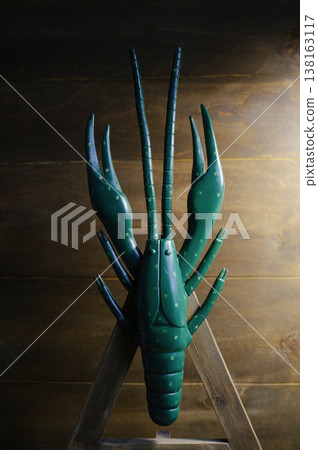 Vertical front view of a large handcrafted wooden crayfish sculpture painted dark green with light green spots, displayed on a wooden easel against a plank wall. Vertical front view of a large handcrafted wooden crayfish sculpture painted dark green with light green spots, displayed on a wooden easel against a plank wall. 138163117