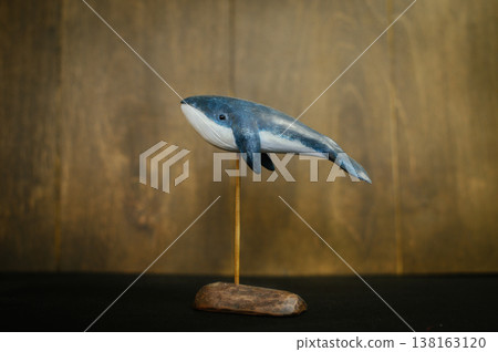 Horizontal side view of a handcrafted wooden blue whale sculpture, featuring a white underbelly and displayed on a thin vertical stand with a rustic base. Horizontal side view of a handcrafted wooden blue whale sculpture, featuring a white underbelly and displayed on a thin vertical stand with a rustic base. 138163120