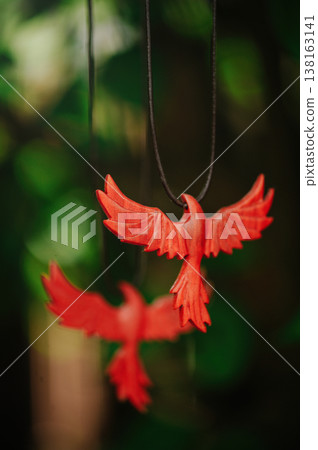 Vertical artistic view of two handcrafted red wooden phoenix pendants, with one in sharp focus and another blurred in the background, against green leaves. Vertical artistic view of two handcrafted red wooden phoenix pendants, with one in sharp focus and another blurred in the background, against green leaves. 138163141