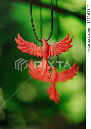 Vertical artistic view of two handcrafted red wooden phoenix pendants, with one in sharp focus and another blurred in the background, against green leaves. 138163142