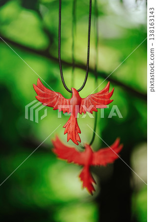 Vertical artistic view of two handcrafted red wooden phoenix pendants, with one in sharp focus and another blurred in the background, against green leaves. Vertical artistic view of two handcrafted red wooden phoenix pendants, with one in sharp focus and another blurred in the background, against green leaves. 138163143