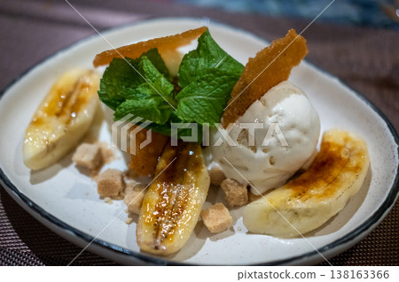 Caramelized banana dessert served with vanilla ice cream, mint leaves, and crispy sugar garnish on a plate. Caramelized banana dessert served with vanilla ice cream, mint leaves, and crispy sugar garnish on a plate. 138163366