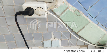 White double electrical outlet is installed on a tiled wall, connected to black and white power cables. The surrounding square tiles show signs of wear, age, and cracked grout White double electrical outlet is installed on a tiled wall, connected to black and white power cables. The surrounding square tiles show signs of wear, age, and cracked grout 138163813