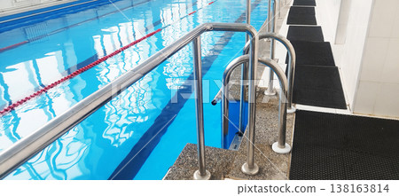 Shiny chrome handrails lead into bright indoor swimming pool. The turquoise water ripples under artificial lights, featuring red lane dividers and black anti-slip mats along the tiled deck for safety Shiny chrome handrails lead into bright indoor swimming pool. The turquoise water ripples under artificial lights, featuring red lane dividers and black anti-slip mats along the tiled deck for safety 138163814