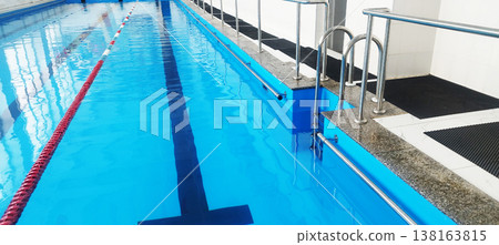 A clean indoor swimming pool features vibrant blue water and red lane dividers. Chrome handrails lead into the water along the stone edge, while black anti-slip mats line the tiled walkway for safety A clean indoor swimming pool features vibrant blue water and red lane dividers. Chrome handrails lead into the water along the stone edge, while black anti-slip mats line the tiled walkway for safety 138163815