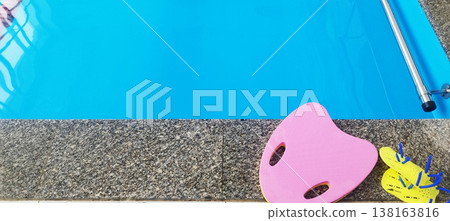 Bright blue pool water is bordered by a speckled grey granite edge. A pink kickboard and yellow hand paddles rest on the deck, prepared for a swim workout or training session at the indoor facility Bright blue pool water is bordered by a speckled grey granite edge. A pink kickboard and yellow hand paddles rest on the deck, prepared for a swim workout or training session at the indoor facility 138163816