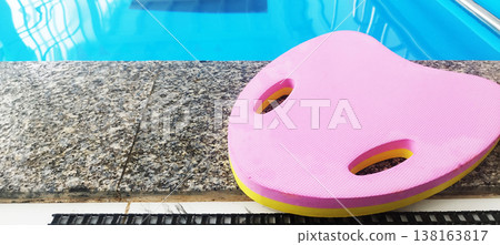 Bright blue water in a swimming pool is edged with a gray granite rim with inlays. A pink kickboard rests on the edge, ready for swimming practice in an indoor pool. 138163817