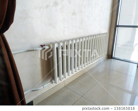 Classic white sectional radiator is mounted on a light-colored textured wall in a bright room. The sunlight from a nearby window reflects off the polished floor tiles, highlighting the heating system Classic white sectional radiator is mounted on a light-colored textured wall in a bright room. The sunlight from a nearby window reflects off the polished floor tiles, highlighting the heating system 138163818
