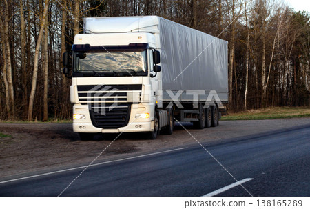 White semi-truck DAF XF with gray curtainside trailer parked on dirt shoulder alongside asphalt highway by bare deciduous forest at dusk or dawn White semi-truck DAF XF with gray curtainside trailer parked on dirt shoulder alongside asphalt highway by bare deciduous forest at dusk or dawn 138165289