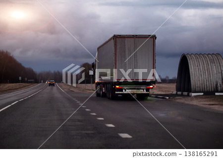 Rear view of large semi-truck traveling on asphalt highway road during overcast day, long distance transport for goods and freight. Copy space for text Rear view of large semi-truck traveling on asphalt highway road during overcast day, long distance transport for goods and freight. Copy space for text 138165291