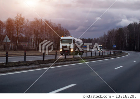 Tanker truck with illuminated headlights travels on curved highway through barren landscape under cloudy evening sky, another truck in distance, evening sunset. Copy space for text Tanker truck with illuminated headlights travels on curved highway through barren landscape under cloudy evening sky, another truck in distance, evening sunset. Copy space for text 138165293