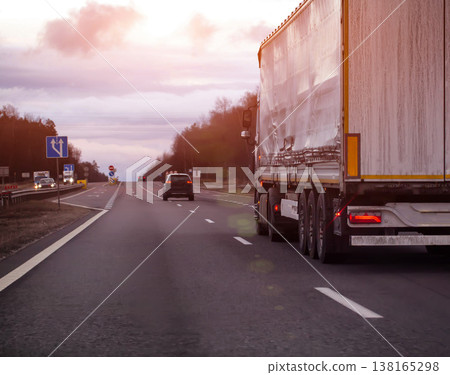 Semi-truck with white trailer traveling on highway at dusk during overcast day, freight transportation along road infrastructure, evening sunset. Copy space for text Semi-truck with white trailer traveling on highway at dusk during overcast day, freight transportation along road infrastructure, evening sunset. Copy space for text 138165298