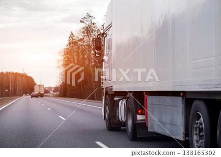 White semi-trailer truck travels highway, side view trailer, wheels, forest, overcast sky, depicting cargo transport logistics industry efficiency, evening sunset. Copy space for text 138165302
